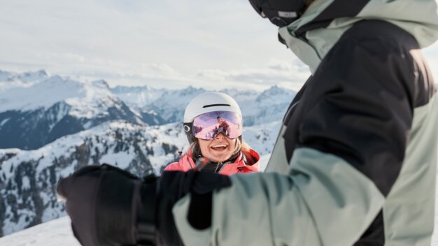 Zwei Skifahrer auf einem verschneiten Berg; im Vordergrund eine Person in grüner Jacke, im Hintergrund eine lächelnde Frau mit weißem Helm und rosa Skibrille vor alpiner Bergkulisse. | © INTERSPORT International Corporation GmbH