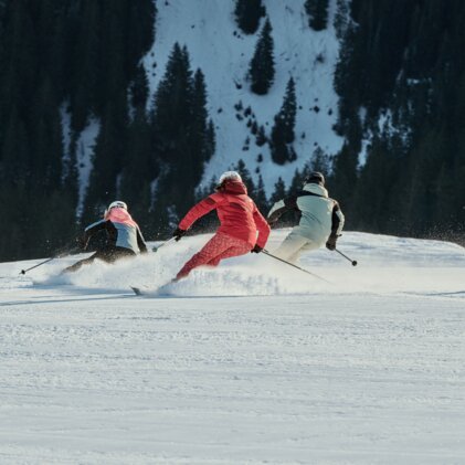 Three skiers in colorful jackets carve parallel turns on a groomed slope, with a pine forest and snow-covered mountainside in the background. | © INTERSPORT International Corporation GmbH