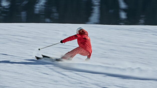 Person in red ski jacket and light-colored helmet takes a fast turn on a snowy slope, snow sprays, dark forest in the background. | © INTERSPORT International Corporation GmbH