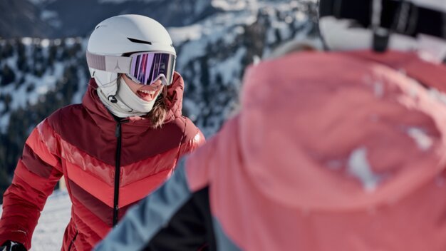 Smiling skier wearing a white helmet and pink ski goggles in a red jacket; blurred companion in the foreground and snowy mountain landscape in the background. | © INTERSPORT International Corporation GmbH
