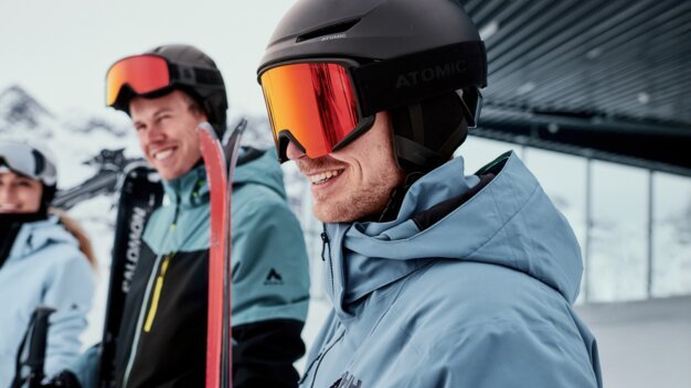 Three skiers wearing helmets and ski goggles on a snowy mountain slope, with a smiling man in a blue jacket in the foreground. | © INTERSPORT International Corporation GmbH