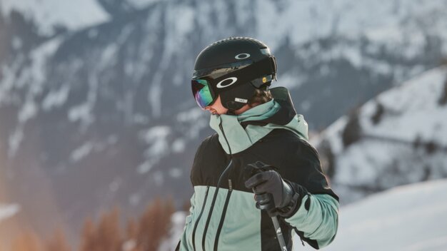 OTG ski goggles Skier in front of skier in black helmet with mirrored Oakley ski goggles and mint-black jacket, holding a ski pole with gloves in front of a snowy mountain landscape. | © INTERSPORT International Corporation GmbH