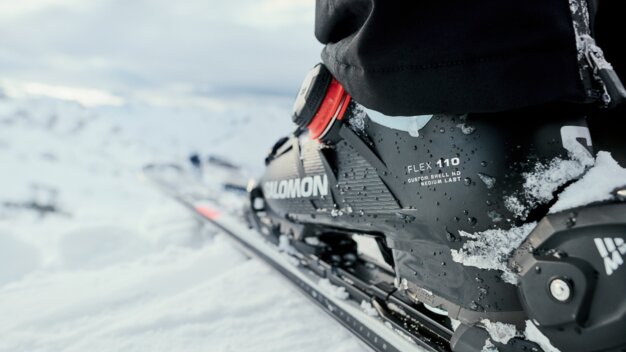 Close-up of a black Salomon ski boot (Flex 110) with snow on a ski binding, person in black ski pants, snowy mountain landscape in the background. | © INTERSPORT International Corporation GmbH