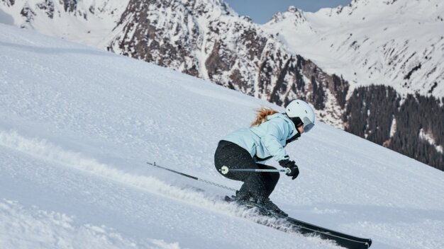 Woman skiing in a light blue jacket and white helmet, she is skiing down a steep, snow-covered slope in front of snow-covered mountains. | © INTERSPORT International Corporation GmbH