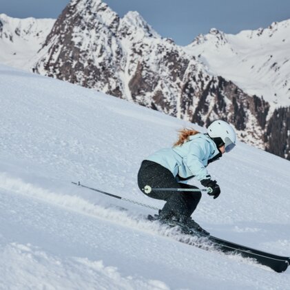 Woman skiing in a light blue jacket and white helmet, she is skiing down a steep, snow-covered slope in front of snow-covered mountains. | © INTERSPORT International Corporation GmbH