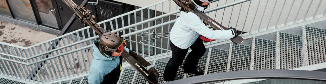 Two people wearing helmets and winter jackets carry skis over their shoulders and climb a metal outdoor staircase, view from above, urban environment. | © INTERSPORT International Corporation GmbH