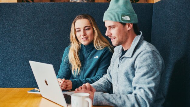 Two people are sitting at a wooden table in a dark alcove, looking at an open laptop; both are wearing warm fleece jackets (The North Face) and there is a coffee cup on the table. | © INTERSPORT International Corporation GmbH