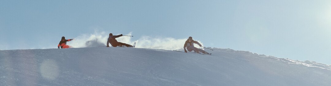 Three skiers descend a wide, snow-covered slope in the sunlight, powder snow spraying behind them. | © INTERSPORT International Corporation GmbH