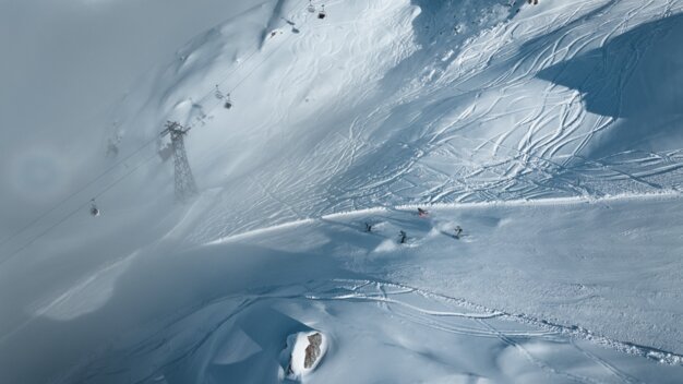 Bird's-eye view: snow-covered mountainside with gondola lift, numerous ski tracks, and four skiers (one wearing a red jacket) | © INTERSPORT International Corporation GmbH