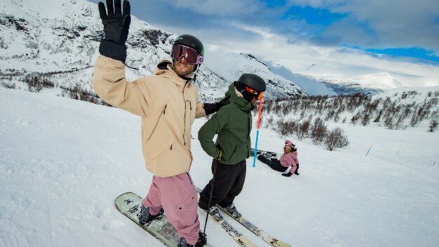 A man on a snowboard stands on the slope next to a skier and raises his hand to wave.