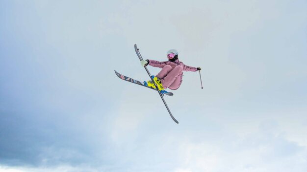 Skiers in pink ski suits with helmets and pink ski goggles perform a jump with their skis crossed against a bright winter sky. | © INTERSPORT International Corporation GmbH