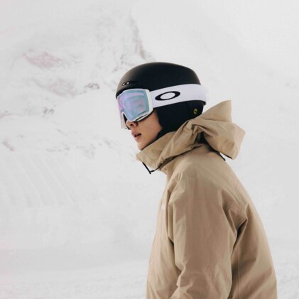 Person im Profil mit schwarzem Skihelm und weißen Oakley‑Skibrille, beige Wintersportjacke vor verschneiter Bergkulisse | © INTERSPORT International Corporation GmbH