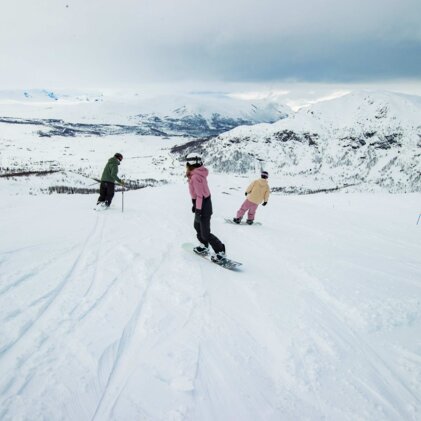 Three people snowboarding on a wide, snow-covered slope with snow-capped mountains in the background and a cloudy sky. | © INTERSPORT International Corporation GmbH