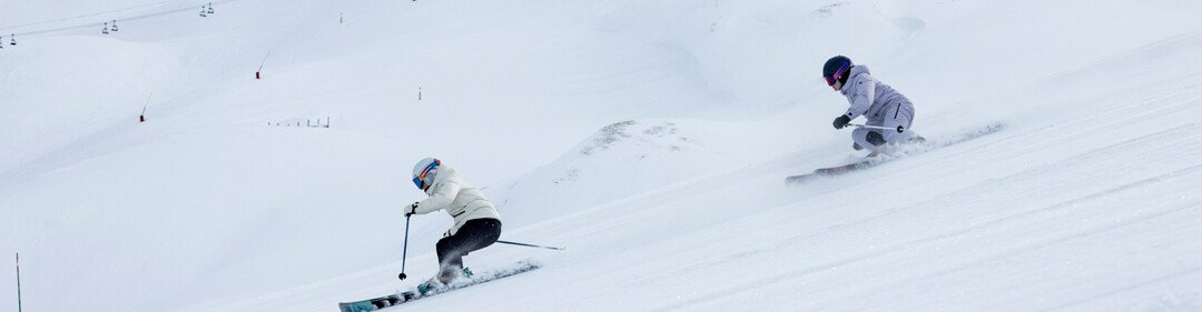 Two winter sports enthusiasts on a snow-covered slope—on the left, a skier in a white jacket with a helmet; on the right, a snowboarder; chairlifts in the background. | © INTERSPORT International Corporation GmbH