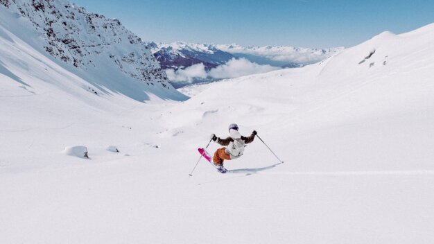 Skifahrende in weißer Jacke und orangefarbener Hose mit pinken Skiern fährt im Tiefschnee vor weitem, verschneitem Bergtal | © INTERSPORT International Corporation GmbH