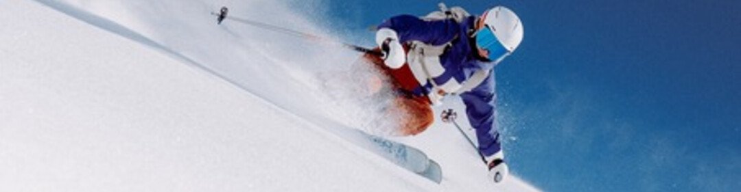 Skiers in blue jackets and white helmets ski diagonally down a steep powder snow slope, snow dust flying everywhere. | © INTERSPORT International Corporation GmbH