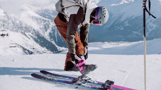 Person in Skihelm und Rucksack, die auf einer schneebedeckten Bergpiste eine pink-schwarze Skischuh-Schnalle an Bindung befestigt; blicken nach unten, Berge im Hintergrund. | © INTERSPORT International Corporation GmbH