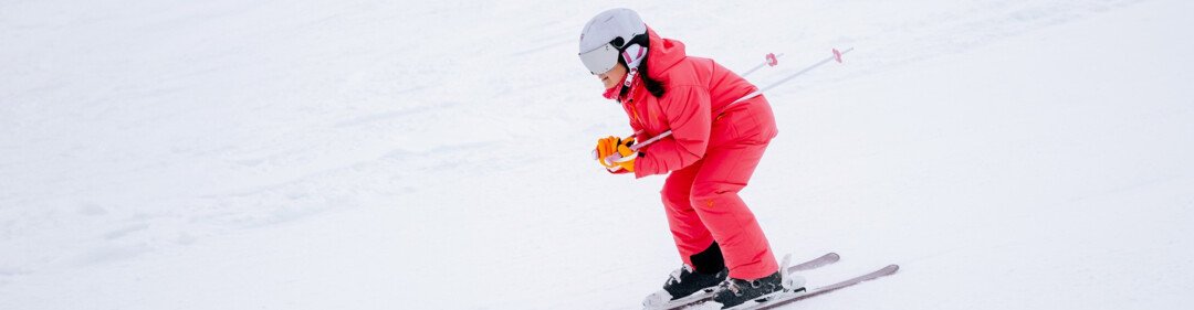 Person in red ski suit and white helmet in a slightly crouched position skiing on a snow-covered slope, wearing orange gloves and carrying ski poles. | © INTERSPORT International Corporation GmbH