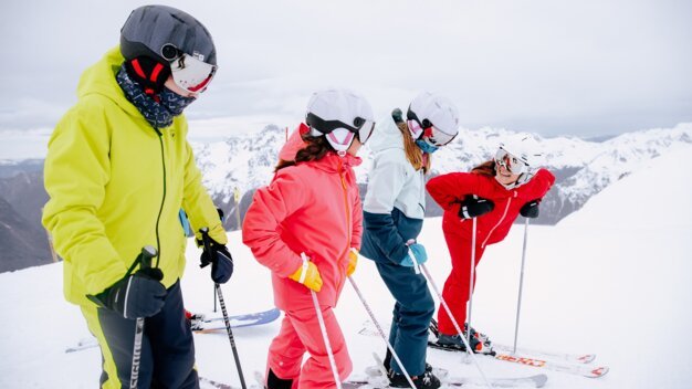Group of four skiers in colorful winter jackets and helmets standing with ski poles at the edge of a snowy mountain slope, snow-covered mountains in the background. | © INTERSPORT International Corporation GmbH