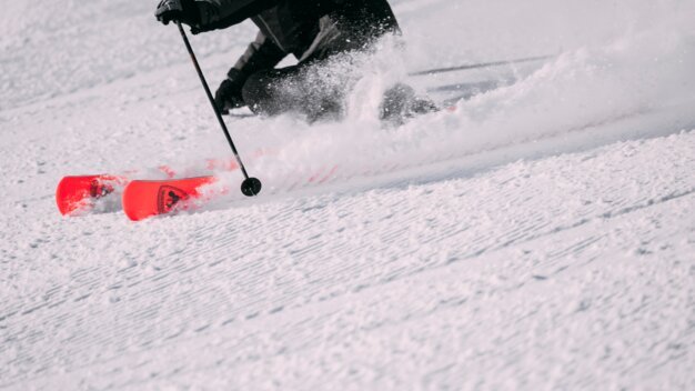 Person carving on skis: lower body and ski poles visible, red Rossignol skis shooting through powder snow and spraying snow. | © INTERSPORT International Corporation GmbH