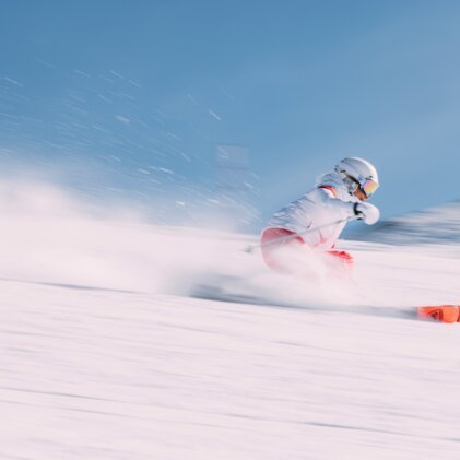Schnell fahrende Skifahrerin in weißer Jacke und roten Hosen mit orangefarbenen Skiern, dynamischer Schneespritzer vor blauem Himmel. | © INTERSPORT International Corporation GmbH