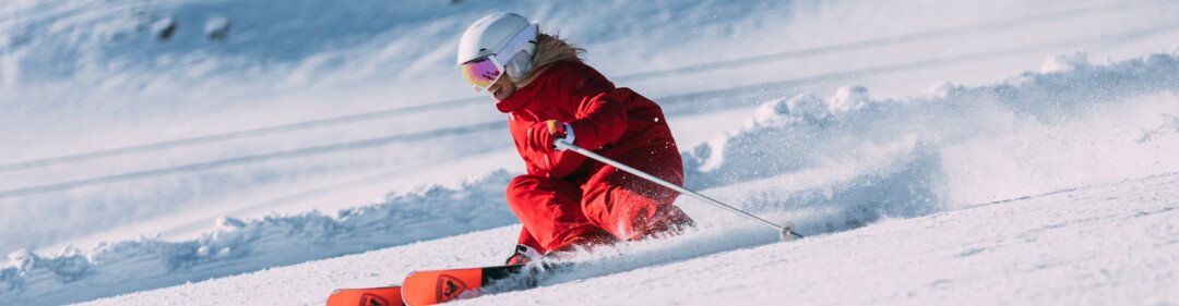 Person in red ski suit and white helmet carving on a sunny, freshly groomed ski slope in front of a snow-covered mountain. | © INTERSPORT International Corporation GmbH