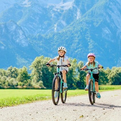 Two kids cycling on a gravel path through a sunny landscape with mountains in the background.