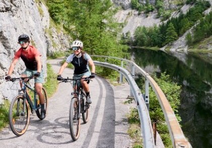 Two cyclists on a narrow path along a river, surrounded by cliffs and lush nature.