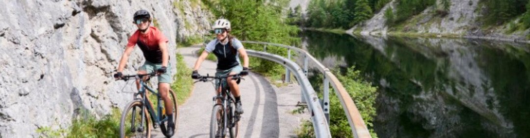 Two cyclists on a narrow path along a river, surrounded by cliffs and lush nature.