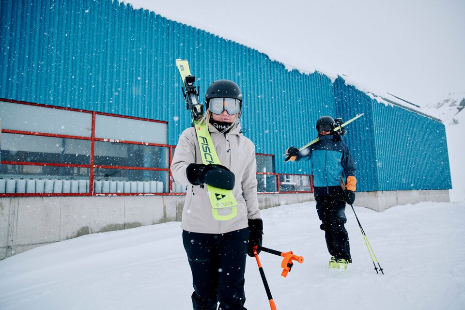 © Fischer Sports GmbH Two skiers with skis in front of a blue building in the snow. | © Fischer Sports GmbH