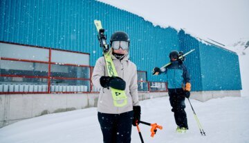 Zwei Skifahrer mit Skiern vor blauem Gebäude im Schnee. | © Fischer Sports GmbH