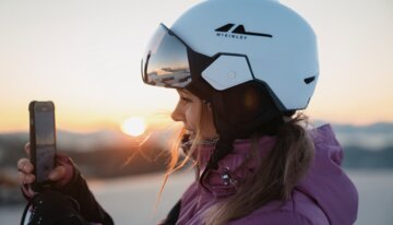 Frau mit Skihelm filmt Sonnenaufgang mit Handy | © INTERSPORT International Corporation GmbH