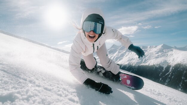 Frau fährt Snowboard auf einer Skipiste und lächelt.