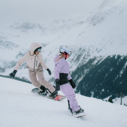 Zwei Snowboarderinnen fahren einen verschneiten Hang hinunter.