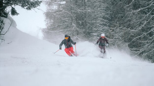 Zwei Skifahrer fahren bei Schneefall einen schneebedeckten Hang hinunter.