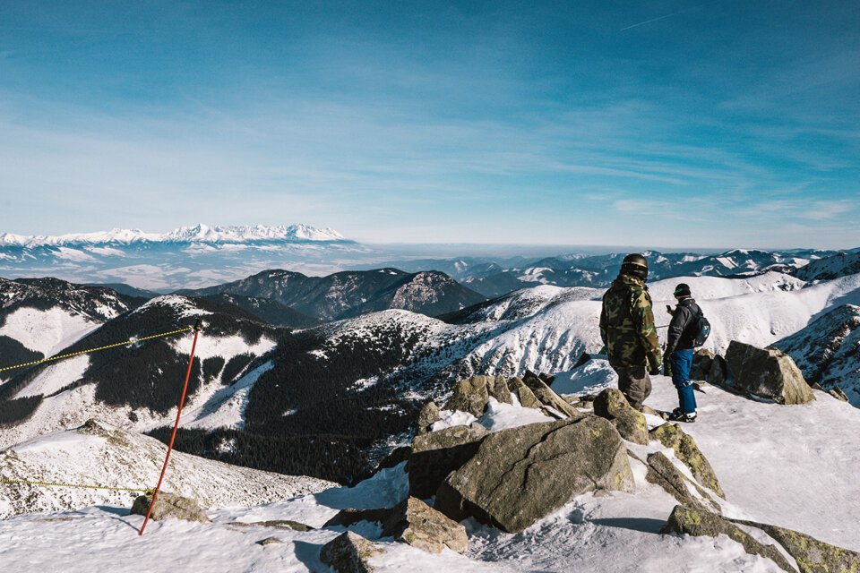 Zwei Personen auf einem schneebedeckten Berggipfel mit Bergpanorama im Hintergrund.