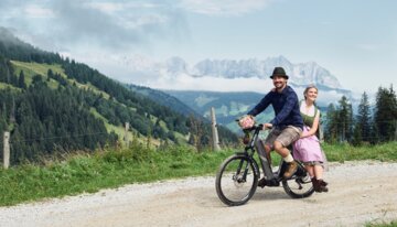 Ein Mann in Lederhose und blauem Hemd fährt auf einem E-Bike, auf dem Gepäckträger sitzt eine Frau in Dirndlkleid. Beide lächeln.
