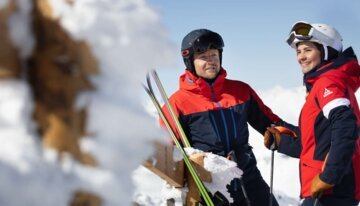 Zwei Skifahrer in Winterkleidung, lehnen an einem verschneiten Holzgeländer.
