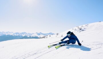 Skifahrerin in Blau fährt eine sonnige, verschneite Piste hinunter.